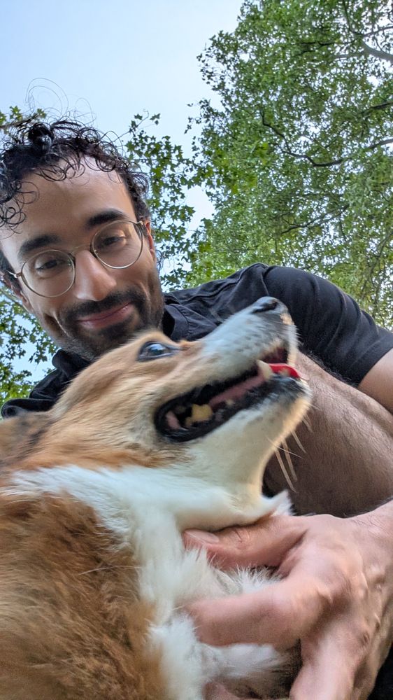 Georgios smiling with a very happy corgi in his arms, under summer trees