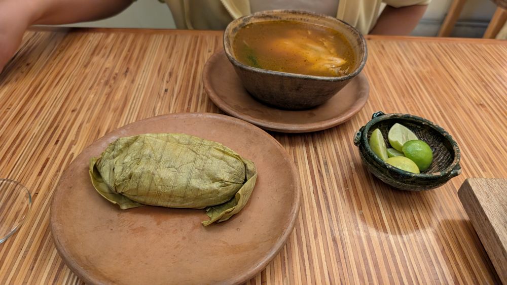 A banana-leaf tamal on a clay plate, beside a bowl of caldo and fresh limes
